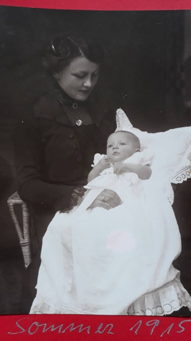 Schwarz/Weiß-Foto. Eine Frau sitzt mit Säugling im Arm auf einem Korbstuhl.